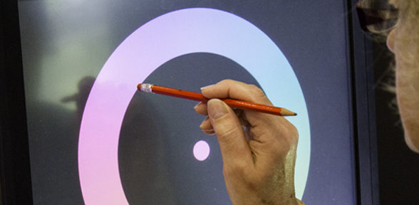 Large touch screen experiment being show with image of an outline gradient and coloured dot in the centre. There is a hand of a person taking part who is using the rubber on a red pencil as a touch input and is about to touch part of the screen.